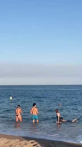 A group of four people are standing in the shallow water of the ocean near a sandy beach. The sky is clear and blue with a faint haze near the horizon. The water is blue and calm with small waves washing ashore. Two people are standing facing away from the camera. One is wearing a black bikini and the other is wearing blue swim trunks. To their right, another person is kneeling in the water, facing away from the camera, wearing a black swimsuit. A fourth person is swimming further out in the ocean, facing the shore. A yellow buoy is visible in the water to the left of the group. The sun is shining, creating a bright scene.