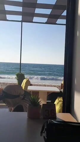 The video shows a view from inside a cafe or restaurant looking out at a beach and the ocean. Outside, there are tables and chairs, some with yellow cushions. The ocean waves are crashing onto the shore. Inside, on a table, there is a small potted plant and a table number sign that reads "36". A black bag is also visible on the table. The lighting suggests it is daytime.