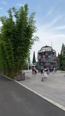 The video shows a walkway next to a lush bamboo grove. A glass structure with colorful panels is visible in the distance. People are walking and cycling on the paved path. The camera pans to the left, revealing a weeping willow tree and a lake filled with lily pads. Boats can be seen on the water. The sky is partly cloudy.