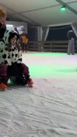 A short video shows a young girl ice skating on an indoor rink. She wears a hooded jacket with a black-and-white cow print, purple pants, and orange skates. At the beginning, she is sitting on a black seat with skate blades, but then she is placed on a white-and-black penguin-shaped skate aid. Someone is pushing the skate aid along the ice. Another child in a red-and-green jacket and a boy in a blue jacket can be seen in the background near a red reindeer-shaped skate aid. The ice rink is decorated with string lights, and the lighting in the rink changes from green to blue to purple.