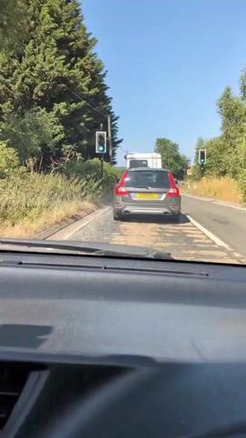 The video is shot from the interior of a vehicle, looking forward through the windshield. The vehicle is on a paved road with a white line marking the lane. In front of the vehicle, there is a grey SUV ahead, and further ahead, a white trailer, possibly a caravan. On the left side of the road, there are tall green trees and bushes. A traffic light with a green signal is visible on a pole on the left side of the road. Another traffic light with a green signal is visible on the right side of the road. The sky is clear and blue, indicating a sunny day. The road appears to have patches of a different texture or color, possibly due to roadwork or wear. The overall scene depicts a slow-moving traffic situation on a rural road.