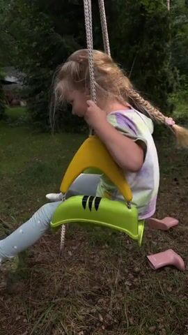A young girl with blonde hair and a tie-dye t-shirt is sitting on a green swing with yellow armrests in an outdoor, wooded setting. She is wearing light blue leggings with a white flower pattern and white socks with a black pattern. Her pink boots are visible on the ground next to the swing. The girl is swinging back and forth and appears to be laughing and having fun. The video is a close-up shot of the girl on the swing.