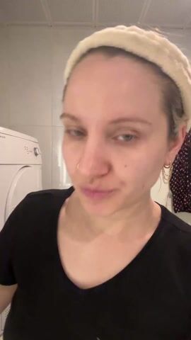 The video shows a woman with blonde hair wearing a white headband and a black t-shirt applying a liquid to her face. She sprays the liquid from a green and white bottle into the palm of her hand and then rubs it onto her face. The background includes a white tiled wall, a white washing machine, and a garment with a heart pattern hanging on the wall.