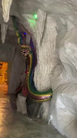 The video pans across the interior of a cave-like structure. Stalactites hang from the ceiling, and the walls are textured to resemble rock formations. Colorful LED lights, green, blue, and yellow, illuminate the space. In the foreground, a large, ornate statue of a multi-headed Naga, a mythical serpent-like creature, is coiled around a rock pillar. The Naga statue is vibrant, with green, gold, red, and blue scales, and a detailed, dragon-like head with red eyes. Further into the cave, an opening reveals a dimly lit chamber with more illuminated yellow structures. As the camera continues to pan, it shows two statues within an alcove. One statue is a dark figure adorned with gold and red, possibly representing a deity, and appears to be seated or standing. Beside it, a lighter-colored statue of a female figure holding a staff is visible. A white, scaled serpent-like object is wrapped around these statues. The floor is a smooth, grey surface.