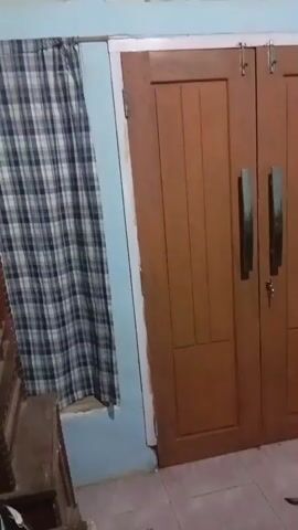 The video shows a dimly lit room with a light blue wall and a wooden door. To the left of the door, there is a window with a blue and white checkered curtain. In front of the door and to the right, there are several motorcycles parked. The motorcycles are of various colors, including black, white, and blue. The floor appears to be tiled. The overall impression is of a storage area or a garage.