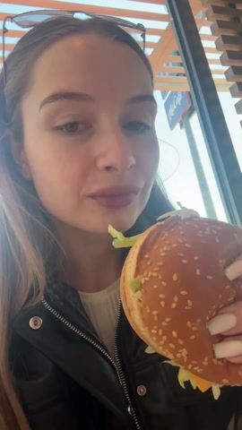 A woman is eating a hamburger in what appears to be a McDonald's restaurant. She is wearing a black leather jacket, a white shirt, and sunglasses are perched on her head. Her nails are long and painted white. The hamburger has a sesame seed bun, lettuce, and cheese. The restaurant's interior features wood slat decorations and a McCafe sign is visible in the background. The woman takes a large bite of the burger, chews, and makes a satisfied expression.
