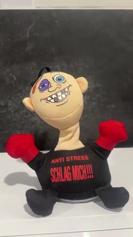 A close-up shot features an anti-stress plush toy against a dark, textured background, sitting on a white surface. The toy has a caricatured face with a fair skin tone, a black eye, a blue eye, black hair, and a wide, toothy grin with missing teeth. Its neck is flexible and spring-like. It wears a black shirt with red text that reads "ANTI STRESS" at the top and "SCHLAG MICH!!!" (German for "HIT ME!!!") below. The toy has red boxing gloves for hands and black feet. A hand with pink painted nails enters the frame and gently taps the toy's head. Upon being touched, the toy emits a high-pitched yelp followed by a lower, guttural sound, reacting to the "hit." The scene implies a humorous interaction with an anti-stress novelty item.