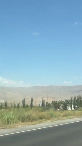 This video appears to be recorded from a moving vehicle on a sunny day. The camera faces forward and slightly to the right, capturing a highway with a white car in front. The landscape features dry, grassy fields on the right side of the road, interspersed with trees and some buildings. In the background, there are large, barren mountains under a clear blue sky with scattered white clouds. There are also power lines visible, indicating a rural or semi-rural setting. The white car in front has a license plate visible, and it appears to be driving away from the camera. There is a road sign with the number 52 on it. The overall impression is of a journey through a dry, mountainous region on a clear day.