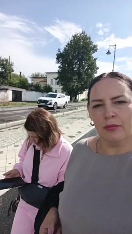 Two women are visible outdoors on a sunny day with a cloudy sky. The woman in the foreground is on the right, facing the camera with a neutral expression. She has dark hair pulled back, is wearing a grey top, and has a necklace on. The woman in the background is on the left, facing away from the camera and looking down at a black smartphone. She has brown hair and is wearing a pink hooded jacket over a black garment, with a black bag slung across her body. A white SUV is parked on a road behind them. The ground is paved with cobblestones and asphalt. There are trees and buildings in the background.
