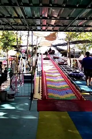 A short video shows a young child on a tube sled going down a colorful, multi-colored slide. The slide appears to be made of a material that resembles small balls. The child is wearing a light-colored shirt and dark pants. The slide is elevated, and there is a roof structure above. Other people can be seen in the background, some standing and some on other slides or structures. The environment appears to be an outdoor amusement park or playground, with trees and other attractions visible.