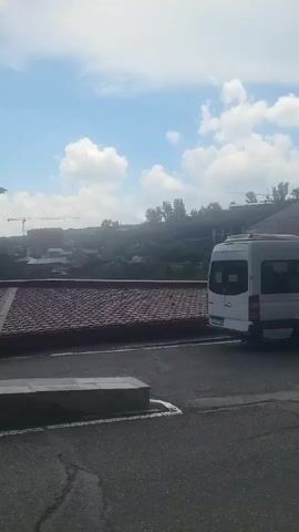 The video pans across an outdoor scene. In the foreground, there is an asphalt surface with white markings. To the left, a portion of a building with a brown, tiled roof is visible. To the right, a white van is parked. Further back, a long building with a red, tiled roof and a glass-enclosed facade stretches across the frame. A red umbrella is positioned near this building. Several people are walking away from the camera on the asphalt. Beyond the buildings, a hilly landscape with trees and some structures is visible under a cloudy sky with blue patches. Construction cranes can be seen in the distance.