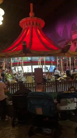 The video begins with a close-up of a brightly lit red carousel. The carousel is decorated in an East Asian style, with ornate carvings and hanging lanterns. Several people are riding the carousel, which is in motion, creating a blur of movement. In the foreground, two strollers are visible, one dark blue and the other white. A person wearing a light pink shirt and black pants stands to the left, looking towards the carousel.

As the video progresses, the camera pans to the left, revealing more of the environment. A wooden pillar with hanging banners and lanterns is visible. More people are milling about in the background, and several food stalls or shops are present. The overall atmosphere appears to be that of an amusement park or entertainment venue at night, with colorful lights and a lively crowd. The camera continues to pan, showing more people walking and interacting, and the carousel remains the central focus of the scene.

Towards the end of the video, the camera pans further to the right, revealing more of the park's architecture and decorations. The carousel continues to spin in the background. The video ends with a view of the park from a slightly different angle, with more people and attractions visible. The lighting is dim but festive, with a mix of artificial lights and what appear to be stage lights. The environment seems to be indoors, possibly a themed indoor amusement park.