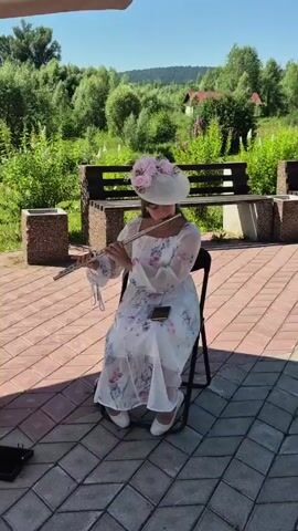 A girl in a white dress and a wide-brimmed hat decorated with pink flowers sits on a black folding chair and plays the flute. She is outdoors on a paved area with a pattern of grey and reddish-brown tiles. Behind her is a wooden park bench with a dark wood top and concrete legs. Beyond the bench, lush green trees and bushes are visible, with a few pink flowers growing near the bench. In the background, a gentle slope covered with trees rises to a hill under a clear blue sky. Sunlight casts shadows on the tiled ground. A black object, possibly a music stand or case, sits on the ground to the left of the girl. A dark object, likely a smartphone, rests on her lap.