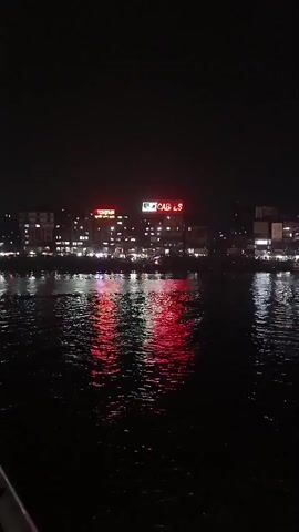 The video shows a nighttime view of a river with city lights reflecting on the water. In the distance, a sign that reads "CABLES" is visible. As the video progresses, two large, brightly lit boats come into view. The boats appear to be stationary and are located near the shore. The water is dark and rippling, reflecting the various lights. The overall scene is dimly lit due to the darkness of the night.