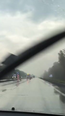 A POV video from inside a vehicle traveling on a wet highway during daylight hours. Raindrops are visible on the windshield. The highway is wet, and there are visible puddles. Traffic cones and construction signs are present on the side of the road. Trees line the right side of the highway. The sky is overcast and cloudy. The vehicle's windshield wipers are briefly visible.