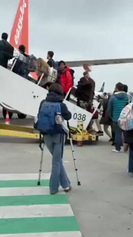 The video shows passengers boarding an EasyJet plane at an airport. A woman with crutches stands at the bottom of the stairs leading up to the plane. The plane is white with an orange tail and the EasyJet logo. Ground equipment, including a stairway and a power unit, are positioned next to the plane. Other passengers are walking up the stairs to board the plane. Another airplane is visible in the background. The weather appears cloudy.