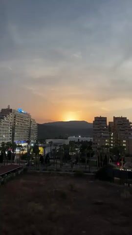 The video shows a sunset over a town with several tall buildings and palm trees. The sky is cloudy with hues of orange, yellow, and gray. The sun is a bright orb peeking over a distant hill. In the foreground, there is dry grass and some shadows. The buildings are illuminated by artificial lights. There are no people visible in the video.