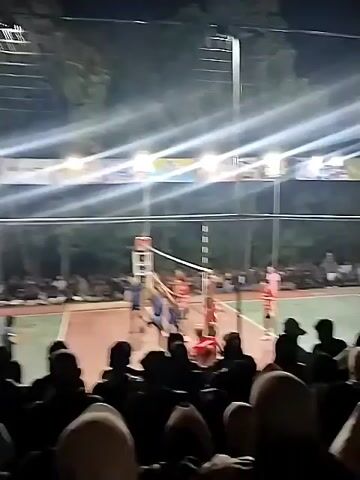 A nighttime volleyball game is taking place outdoors under bright lights. A group of spectators is visible in the foreground, silhouetted against the lights. In the background, players in blue and red uniforms are actively engaged in the game on a volleyball court. The court appears to be made of red material with white lines marking boundaries. A net is stretched across the middle of the court. Trees and foliage are visible behind the court, and banners are hung above the playing area. The overall atmosphere seems to be one of a community event or friendly competition.
