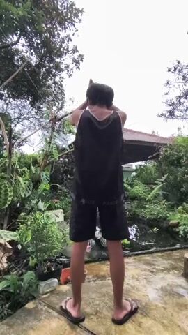 The video shows a person with short dark hair, standing outdoors in what appears to be a garden. They are wearing a black tank top with a white tank top underneath, black shorts with the word "ASICS" visible, and flip-flops. They are standing on a concrete surface with a slightly mottled appearance.

The person is facing away from the camera, with their hands raised above their head. They appear to be making shapes with their hands, perhaps imitating the shape of an animal.

The garden area includes various types of plants, including leafy green plants and some potted plants. In the background, there is a pond or body of water with surrounding greenery. There is also a glimpse of a building with a roof.

The lighting suggests it is daytime, but the sky appears overcast.
