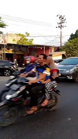 The video shows a street scene with several vehicles and pedestrians. In the foreground, a person wearing a blue hijab and beige cardigan walks across the street, holding a black bag. To the right, a motorcycle is parked. In the middle ground, a gray minivan drives past, and other cars and motorcycles are visible on the road. To the left, a white SUV is parked, and a building with awnings and signage is visible. The environment appears to be an urban or semi-urban area with trees and power lines. The lighting suggests it is late afternoon or early evening.