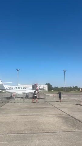 The video shows an outdoor scene with several small airplanes parked on an asphalt surface, likely an airport tarmac. The sky is clear and blue. Several people are present, some standing and observing the airplanes, while others appear to be interacting or moving around. One person is on a scooter, and another is adjusting their hair. There are metal barriers set up in front of some of the airplanes, possibly to keep spectators at a distance. Two lampposts are visible in the background, as well as some low buildings and vegetation. The overall impression is of an event or exhibition involving aircraft.