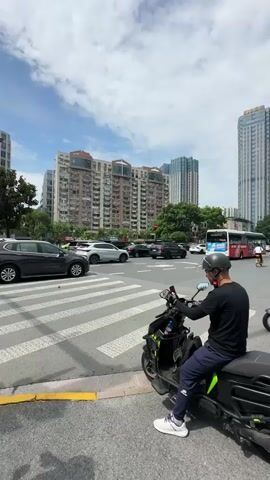 A person wearing a helmet and a black long-sleeved shirt is sitting on a scooter at a crosswalk. The scooter has a black box on the back with stickers on it. In the background, there are several tall buildings, a bus, and cars. The sky is cloudy. The person on the scooter is looking forward and appears to be waiting for the traffic light to change. Another person is standing next to the scooter.