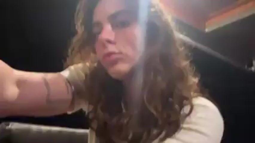 A young woman with long, wavy brown hair is looking directly at the camera. She's wearing a white t-shirt and appears to be standing on a boat or a porch with a roof.  The background is dark, suggesting it's nighttime. There are faint lights in the distance. She briefly touches her hair and puffs out her cheeks. There are voices speaking in the background.