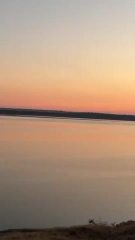 The video shows a wide shot of a body of water with a sunset in the background. The sky is a gradient of colors, from pale blue at the top to orange and pink near the horizon. The water reflects the colors of the sky, creating a soft, warm glow. The foreground is a grassy slope with a few scattered trees and bushes. The scene is peaceful and serene. There are no people or animals visible in the video. The video appears to have been shot from a slightly elevated position, looking out over the water.