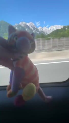 A person's hand holds a colorful stuffed cat in front of a car window. Outside the window, a highway is visible with a concrete barrier and trees lining the road. In the distance, snow-capped mountains are seen under a clear blue sky. The stuffed cat has a striped pattern in orange, pink, yellow, and purple, with yellow eyes. The video is shot from inside a moving vehicle, causing a slight blur in the background.