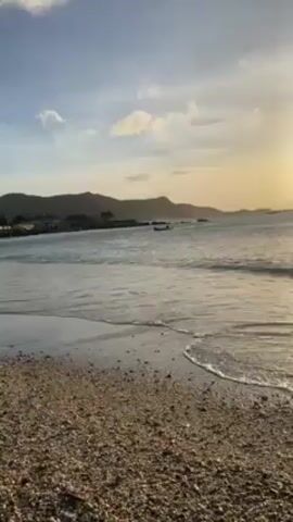 The video shows a beach at sunset. The sun is low in the sky, casting a golden light over the water. Small waves are gently lapping at the shore, which is covered in pebbles. In the distance, there are mountains. There is a small boat in the water.
