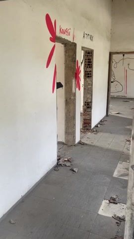 The video captures a walkthrough of an abandoned or neglected building, likely a former school or public institution, characterized by graffiti and dilapidated conditions.

The video begins at 0:00 with a view down a hallway. The walls are white but dirty, and the tiled floor is covered in rubble and debris. There are multiple doorways visible on the left, without doors, framed by concrete. Red, flower-like graffiti adorns the wall around the first doorway. Above the doorways, text graffiti reads "kaussi" with a downward arrow, and "A TYT NOX". Further down the hall on the right, indistinct black graffiti, including the word "MARIA," can be seen. Sunlight streams in from the right, casting shadows.

At 0:01, the camera pans right to a section of a white wall, revealing vibrant, colorful graffiti. The text "LEARN TO BE FREE" is written in a bold, block-letter style using blue, yellow, green, pink, and orange colors. Below this, there's a smaller "23." and a stylized line.

By 0:02, the camera continues to move, showing a concrete staircase leading downwards. On the wall to the left of the stairs, an open, empty, red wall-mounted box (possibly a fire extinguisher cabinet) is visible. More faint scrawls and graffiti are present on the walls.

From 0:03 to 0:04, the camera enters a larger room that appears to be a lecture hall or classroom. Several rows of old, wooden chairs with metal frames are visible, some overturned or in disarray, and all covered in dust. The floor is concrete and dirty. Two large windows on the right wall allow bright natural light into the room. The ceiling is constructed from wooden planks. On the right wall, below the windows, there is blue graffiti, including a large, stylized letter "S" and other shapes. The walls are generally white with signs of wear and tear. The room appears deserted and quiet.