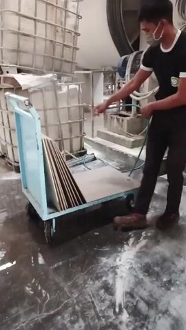 A young man wearing a black t-shirt, dark pants, and a mask sprays a light blue cart filled with flat, rectangular objects. The cart has large, square containers behind it. The man is holding a blue hose and a spray nozzle, and a mist of liquid is coming from the nozzle. The floor is wet and appears to have white powder on it. The background shows industrial equipment, including large tanks and machinery. The man moves the cart, and the spray continues.