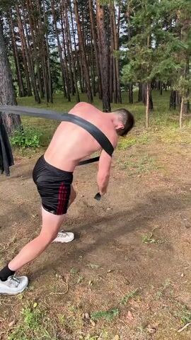 A young, shirtless Caucasian male with short brown hair is performing a resistance band exercise outdoors in a forest. He is wearing black athletic shorts with red stripes and white sneakers. The male is gripping the ends of a black resistance band, which is looped around his upper back. He is bent at the waist and appears to be pulling the band forward and outward, engaging his back and arm muscles. The environment consists of dirt ground, sparse grass, and numerous tall pine trees with sunlight filtering through the branches. A black piece of fabric, possibly clothing, is hanging from a tree trunk to the left of the frame. The video captures several repetitions of the movement.