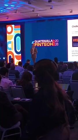 A man in a suit is giving a presentation on a stage in a large room. He is standing in front of a large screen that says "GUATEMALA FINTECH 2026" and has the title "Challenges". The room is filled with people sitting in chairs and watching the presentation. Some of the people are looking at laptops. The room is well-lit and has a modern look. The stage has blue and purple lighting. The audience is mainly men wearing business attire.
