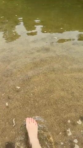 The video shows a body of water with a sandy bottom. The water is clear enough to see the sand and some small debris. Trees and structures are visible on the far bank, and a person's finger is briefly visible in the foreground. The overall scene appears to be a natural or semi-natural water environment, possibly a lake or slow-moving river, with some human elements in the background.