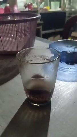 A close-up shot focuses on a glass with a dark liquid residue at the bottom, placed on a light-colored, textured surface. To the right of the glass, a blue bowl with a geometric pattern sits, casting a distinct shadow. In the background, a pink mesh basket is partially visible on the left. The environment appears to be indoors, possibly a room or outdoor seating area, with furniture and shelves faintly discernible in the blurred background. The lighting suggests it might be evening or dimly lit.
