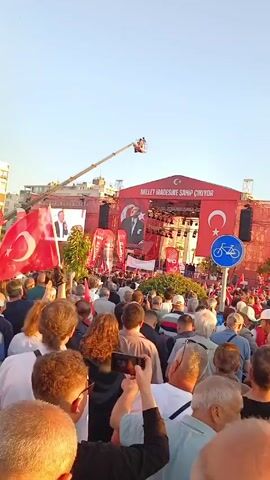 Here is a description of the video:

The video shows a large crowd of people gathered in an outdoor plaza, likely for a political rally or demonstration. The crowd is facing a stage decorated with banners featuring Turkish flags and a portrait of Mustafa Kemal Atatürk. Many people in the crowd are waving Turkish flags. A crane with a platform is visible above the crowd. Buildings surround the plaza, suggesting an urban setting. The sky is clear and the sunlight suggests that it is either early morning or late afternoon. The crowd appears to be listening to someone speaking from the stage, as people are facing the same direction. Several people are using their phones to record or take pictures of the event.