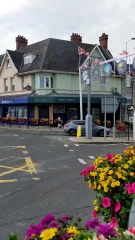 The video shows a street scene with a road, sidewalk, and buildings. There are several cars visible, including a silver SUV and a blue sedan. The sidewalk is lined with black metal railings and planters filled with colorful flowers, primarily pink and yellow petunias. A large archway spans the street, adorned with flags and circular images. Flags, including the Union Jack and what appears to be the Ulster Banner, are prominently displayed. One building has a sign that reads "Woman's Home & Lifestyle." A person is walking on the sidewalk in front of this building. The sky is overcast.