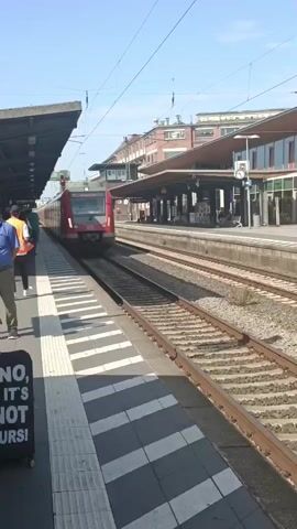 A red train approaches a station platform on a sunny day. The train has the DB logo. A person in a high-visibility vest stands on the platform. There are buildings and overhead wires visible in the background. The platform has a black and white checkered pattern. The train has the RMV logo and a wifi symbol.