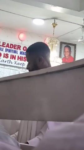Here's a description of the video:

The video shows the head and shoulders of a man standing behind what appears to be a partition or railing, possibly in a church or similar setting. He has short, dark hair and is wearing a white garment.

In the background, there is a sign that reads, "LEADER O. SOLE SPIRITUAL HEAD OF IS IN HIS EARTH KEEP SILEN," though the full text is partially obscured. Above the sign, there's a light fixture and a framed picture of a man wearing a red top. Additionally, a chandelier hangs from the ceiling. Other people in white garments are also present, but they are out of focus.