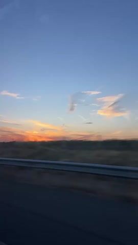 This video captures a sunset viewed from a moving vehicle. The sky is a gradient of blue, transitioning to orange and yellow near the horizon where the sun is setting. Wispy clouds are scattered across the sky. A dark, blurred line of vegetation or land separates the sky from the foreground. In the immediate foreground, there is a blurred, dark road surface and a light-colored guardrail that runs horizontally across the frame. The motion blur indicates that the camera is in transit, likely on a highway.