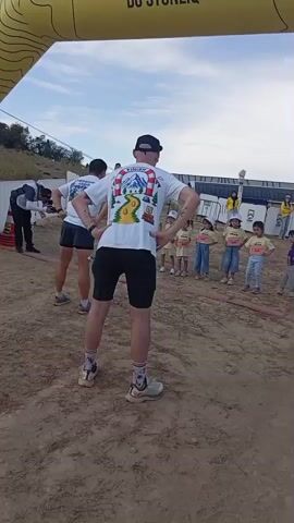 A group of people, including children, are gathered outdoors under a bright sky. A large yellow archway with text on it spans across the top of the frame. The ground is a sandy, uneven surface. In the foreground, a person wearing a white t-shirt with a graphic on the back and black shorts stands facing away from the camera. Their hands are on their hips. To the left, another person in a similar t-shirt and black shorts is also standing with hands on hips. In the middle ground, a group of children, all wearing yellow t-shirts, are lined up facing the person in the foreground. Some adults are visible in the background, including one in a white shirt and black pants to the left of the frame. There are structures and a fence in the background. The overall impression is an outdoor event, possibly a race or gathering for children.