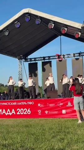 A group of about a dozen dancers are performing on an outdoor stage, silhouetted by bright afternoon sunlight. They’re wearing coordinated outfits: white shirts, dark pants, and dark shoes. They begin the routine in a crouch, then rise to a standing position, engaging in a series of coordinated movements. The stage has a black roof structure supported by a scaffolding frame and multiple stage lights. Large black vertical screens are positioned behind the dancers. The front of the stage is marked with a red banner with white text “ТУРИСТСКИЙ ЛАГЕРЬ ПФО”. Logos are placed along the banner, including the symbol of the Russian Federation and the “ФОСАГРО” logo. The stage sits on a grassy field. A young woman in a red shirt and denim shorts stands in the foreground, partially obstructing the view. People stand in the background, beyond the grassy field.