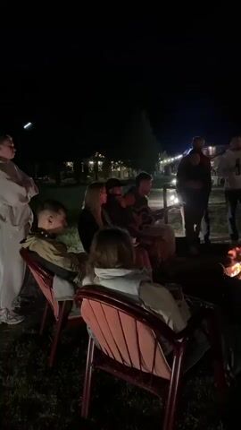 A group of people are gathered around a campfire at night. Some are sitting in chairs, while others are standing. One person is playing a guitar. The fire is burning brightly, casting a warm glow on the faces of the people around it. In the background, there are wooden cabins and string lights. The overall atmosphere appears to be relaxed and social.