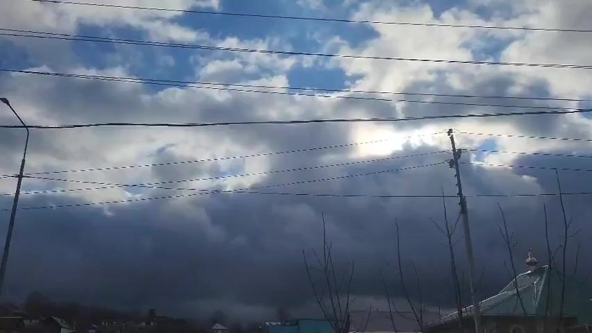 The video presents a static outdoor scene during the daytime, focusing on a dramatic sky above what appears to be a town or village. The sky is divided by contrasting weather conditions: the upper portion reveals patches of bright blue sky with scattered white clouds, while the lower and mid-sections are dominated by large, dark grey, heavy clouds, suggesting an approaching storm or significant cloud cover. The sun is visible on the right side of the frame, peeking through a break in these darker clouds, creating a bright spot of light.

Multiple parallel power lines stretch across the frame, supported by a street light pole on the far left and another utility pole on the right. Below the power lines, the tops of bare tree branches are visible, indicating either winter or early spring. On the far right, the distinctive golden dome of a building, likely a church, can be seen. No people or specific activities are visible in the shot. There is no discernible text. The scene conveys a sense of dynamic weather, with the interplay of sunlight and dark clouds.