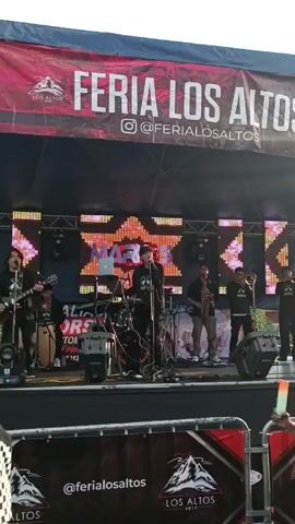 The video shows a band performing on an outdoor stage at an event called “Feria Los Altos”. There are at least six band members playing instruments, including guitar, drums, saxophone, and trombone. The band appears to be singing and playing a lively song. The stage has LED screens behind the band displaying colorful and changing patterns. A banner above the stage advertises the event and its Instagram handle. In front of the stage, a small crowd is gathered, including a woman with blonde hair wearing a leopard print outfit and sunglasses. She is carrying items in her hand and moves around, suggesting she may be leaving or finding a better spot. The environment seems to be a fair or festival during the day, with a partly cloudy sky visible in the background.