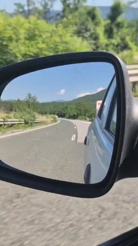The video is a first-person perspective, showing a view of a road and surrounding scenery through a car's side mirror. The mirror reflects a two-lane road curving through a hilly, wooded area. The sky is blue with some clouds, and the sun appears to be shining. Trees and greenery line the sides of the road. The car itself is partially visible in the right side of the mirror. A white car is in front of the reflective car.