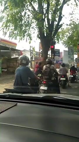 The video shows a street scene with several motorcycles and a truck. The perspective is from inside a vehicle, likely a car, looking forward. In the foreground, the dashboard and windshield of the car are visible. A red traffic light is visible in the distance, indicating that traffic is stopped. Several people are riding motorcycles, most wearing helmets. One motorcycle in the foreground has a person with a backpack. Another motorcycle has two people, one in a striped shirt and one in a white top. A large truck is stopped in the middle of the frame. Trees and buildings line the street, and the sky is bright, suggesting daylight. The overall impression is of a busy street intersection with traffic congestion.