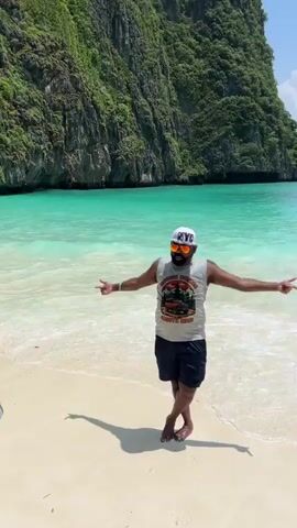 A person wearing a white baseball cap and sunglasses poses for a photo on a beach. They are wearing a grey sleeveless t-shirt with a graphic and dark shorts. The person has their arms outstretched and is looking at the camera. They are standing in shallow water with waves lapping around their feet. In the background, there are large, green, rocky cliffs and clear turquoise water. Another person is visible in the foreground, holding up a black smartphone to take a picture of the person posing. This second person is wearing a white cap and a striped shirt. The overall environment is a tropical beach with bright daylight.