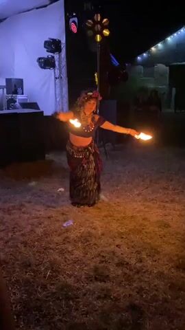 A woman in a dark costume with a skirt and a cropped top is performing a fire dance. She is holding a flaming prop in her right hand and another flaming prop in her left hand. The performance is taking place outdoors at night. There is a stage with lights and speakers in the background. The ground is covered in grass and dirt. There are other people in the background, some of whom appear to be watching the performance. The lighting is dim and appears to be from stage lights and the fire props. The woman is moving dynamically, twirling the flaming props in arcs around her body. Her costume appears to be embellished with beads or metallic elements. A flower or floral accessory is visible in her hair. The overall atmosphere suggests an outdoor event, possibly a festival or performance gathering.